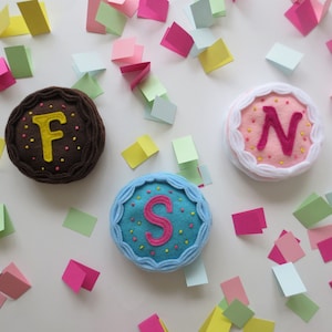 May include: Three felt cakes with different colored frosting and embroidered letters 'F', 'S', and 'N' on top. The cakes are surrounded by confetti on a white background.
