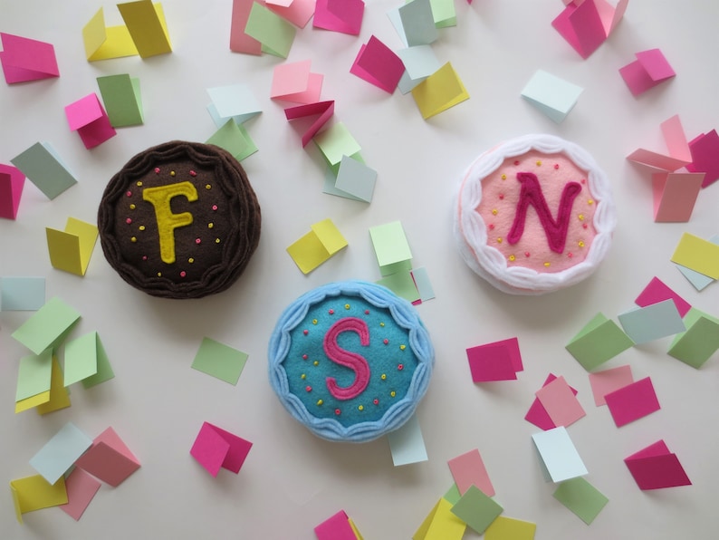 May include: Three felt cakes with different colored frosting and embroidered letters 'F', 'S', and 'N' on top. The cakes are surrounded by confetti on a white background.