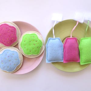 May include: Three round felt biscuits with pink, green, and blue icing and colourful sprinkles on a pink plate, alongside three felt tea bags in light blue, pink, and green with white tags on a light green plate, arranged on a white surface.