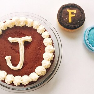 May include: A red velvet cake with white frosting and the letter "J" piped in white on top. There are two small cakes with the letters "F" and "S" on them.