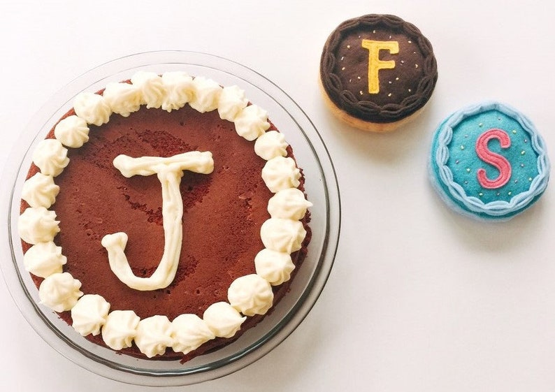 May include: A red velvet cake with white frosting and the letter "J" piped in white on top. There are two small cakes with the letters "F" and "S" on them.