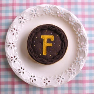 May include: A brown felt cake with a yellow felt letter F on top. The cake is on a white plate with a decorative edge.