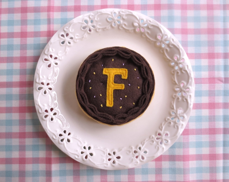 May include: A brown felt cake with a yellow felt letter F on top. The cake is on a white plate with a decorative edge.