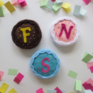 May include: Three felt cakes with different colored frosting and letters F, N, and S embroidered on top. The cakes are surrounded by colorful confetti.