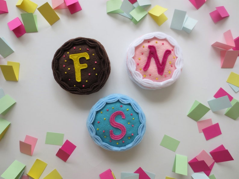 May include: Three felt cakes with different colored frosting and letters F, N, and S embroidered on top. The cakes are surrounded by colorful confetti.