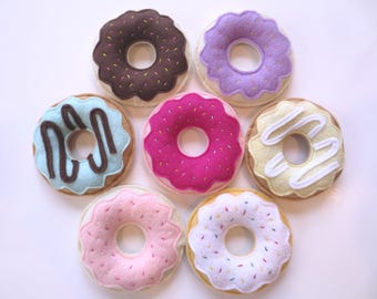 May include: A collection of seven felt donuts arranged in a circular pattern. The donuts feature various colors including brown, purple, light blue, hot pink, pale yellow, light pink, and white, decorated with colorful sprinkles or icing drizzles.