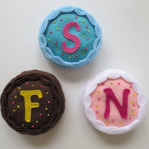 May include: Three felt cake decorations with letters F, N, and S embroidered on them. The cakes are decorated with sprinkles and frosting.