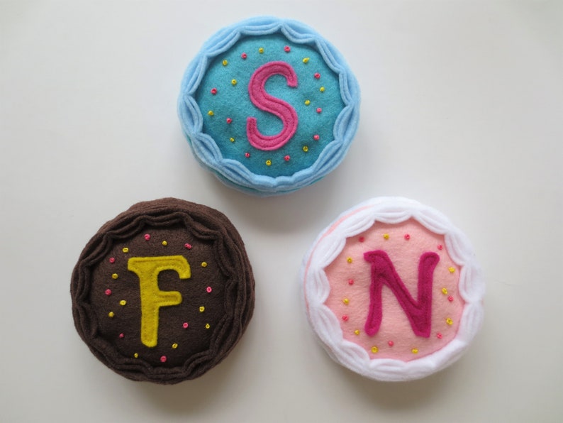 May include: Three felt cake decorations with letters F, N, and S embroidered on them. The cakes are decorated with sprinkles and frosting.
