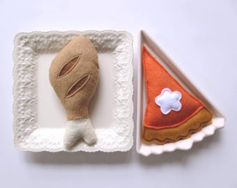 May include: A tan felt chicken leg with two brown slashes and a white bone end on a square white decorative plate, next to a triangular slice of orange felt pumpkin pie with a white flower on a matching triangular white plate.