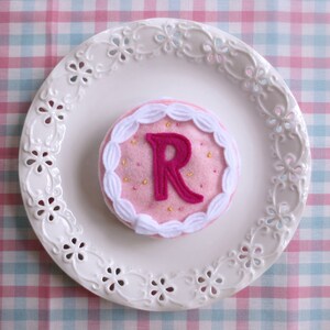 May include: A pink felt cake with white frosting and a pink felt letter R in the center. The cake is on a white plate with a decorative edge.
