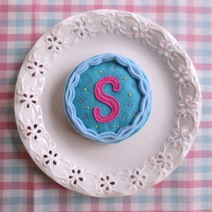 May include: A blue felt cake with a pink felt letter S and yellow sprinkles on a white plate with a decorative edge.