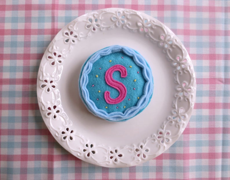 May include: A blue felt cake with a pink felt letter S and yellow sprinkles on a white plate with a decorative edge.