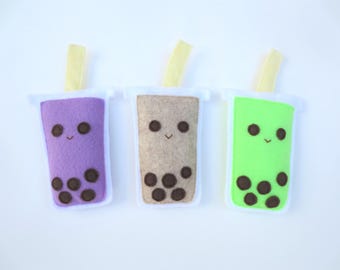 May include: Three felt boba tea plushies in purple, tan, and bright green. Each cup features a white border, a yellow straw, and a stitched face with two circular eyes and a small mouth, along with dark brown circular felt pieces representing tapioca pearls.