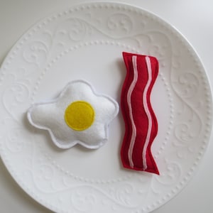 Può includere: Un uovo di feltro bianco con un tuorlo giallo e una striscia di pancetta di feltro rosso su un piatto bianco con un bordo decorativo.