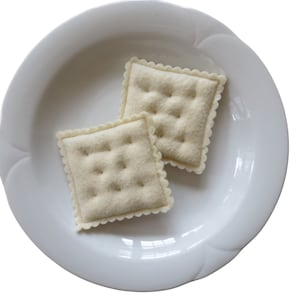 Op de afbeelding: Twee off-white vilten crackers met geschulpte randen en gestikte details liggen in een witte, geschulpte bord. De crackers zijn vierkant met kleine, gelijkmatig verdeelde, gestikte vierkanten.
