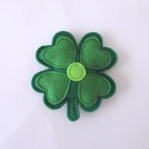 May include: A four-leaf clover ornament crafted from layered green felt. The design features four heart-shaped leaves in a medium green shade, outlined with a darker green border, and a small, circular light green felt accent in the centre.