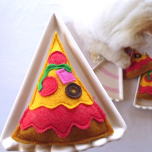 May include: A white cat is playing with a felt pizza toy on a white plate. The pizza toy is yellow with red sauce, green peppers, red and black olives, and pink ham.