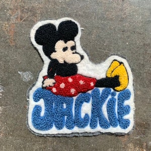 Op de afbeelding: Een witte, blauwe en rode viltpatch met een cartoonafbeelding van Mickey Mouse die een rood-wit geblokt badpak draagt. De patch heeft de naam "JACKIE" in blauwe viltletters.