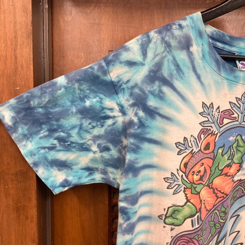 Puede incluir: Una camiseta te&ntilde;ida con un patr&oacute;n de espiral azul y blanco. La camiseta presenta un gr&aacute;fico de un oso con sombrero y bufanda, con copos de nieve y la palabra "RY" en un c&iacute;rculo. La camiseta est&aacute; colgada de una percha negra.