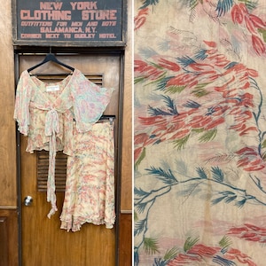 Puede incluir: Un conjunto de dos piezas con estampado floral. La parte superior es una blusa transparente con un lazo en el cuello. La falda es una falda fluida, hasta la rodilla. La tela es de color beige claro con diseños florales rosas, azules y verdes.