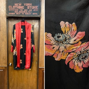 Puede incluir: Una bata de seda negra con forro de satén rojo y estampado floral. La bata tiene cuello chal y mangas largas. El estampado floral presenta flores grandes y estilizadas en tonos de rosa, naranja y amarillo.
