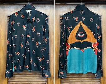 May include: A black long-sleeved shirt with a geometric pattern on the front and a large eye design on the back. The eye is surrounded by two hands in a prayer position.