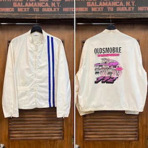 May include: A white jacket with blue racing stripes on the left side. The back of the jacket has a graphic of a pink Oldsmobile car with the text "OLDSMOBILE Takes the WORRY out... of BEING CLOSE!!" and "772" printed on it.