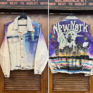 May include: A denim jacket with a tie-dye effect in shades of blue, purple, and white. The jacket features a painted image of the Statue of Liberty and the New York City skyline with the words "New York" in rhinestones.