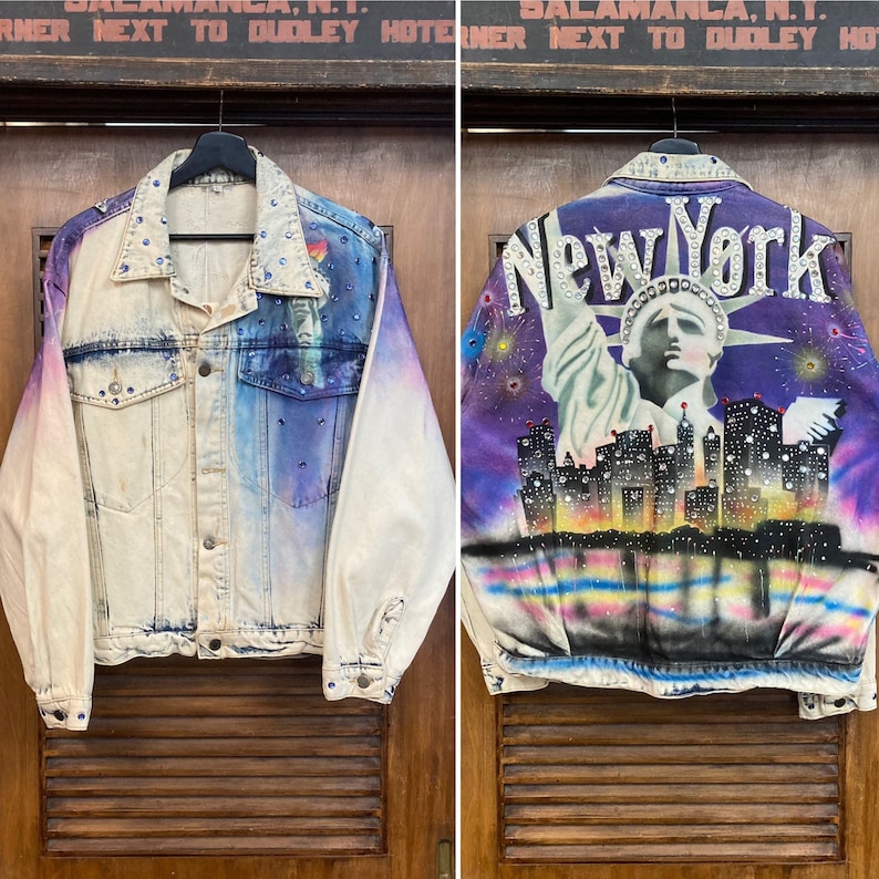 May include: A denim jacket with a tie-dye effect in shades of blue, purple, and white. The jacket features a painted image of the Statue of Liberty and the New York City skyline with the words "New York" in rhinestones.