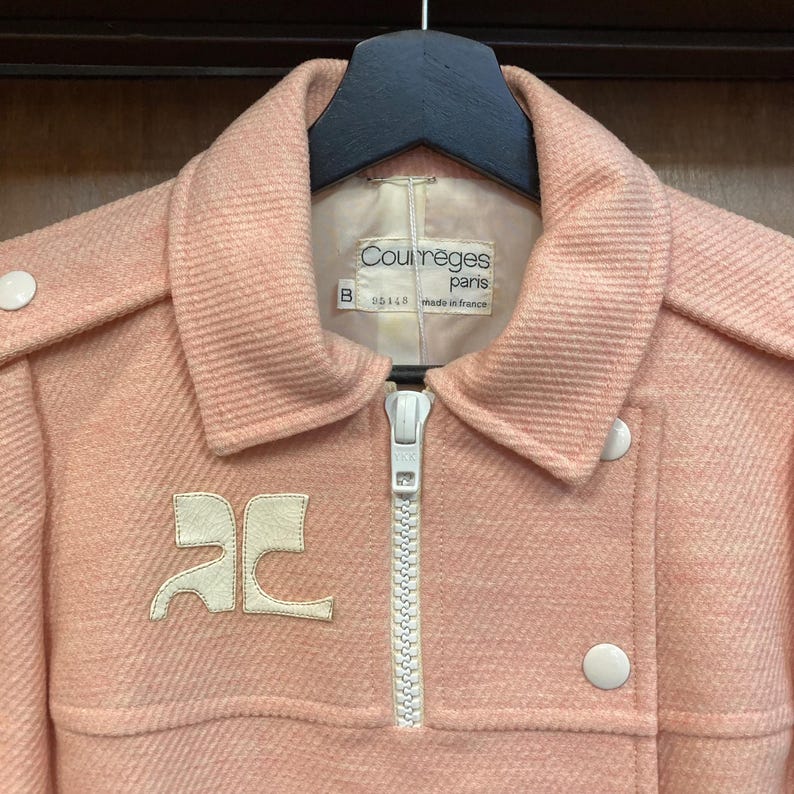 May include: A pink wool coat with a white zipper and white buttons. The coat has a white logo on the front that says "Courreges Paris" and "made in France".