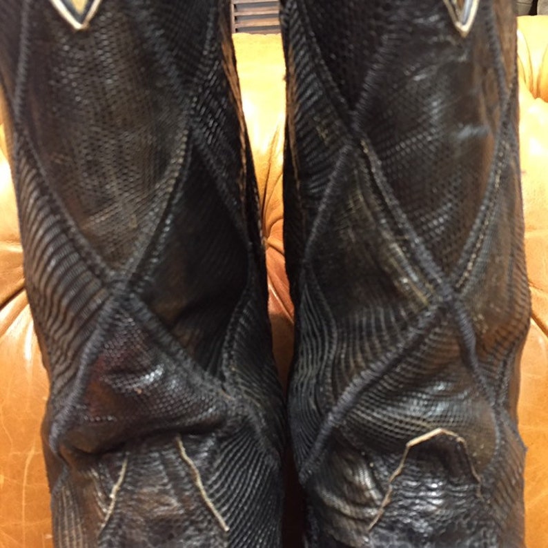 Pu&ograve; includere: Un paio di stivali da cowboy in pelle nera con un motivo a forma di diamante. Gli stivali hanno una punta a punta e un tacco alto.