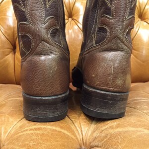 K&ouml;nnte beinhalten: Ein Paar braune Cowboy-Stiefel aus Leder mit spitzer Zehenpartie und einem gestickten Design am Schaft. Die Stiefel haben eine dunkelbraune Sohle und einen leichten Absatz.