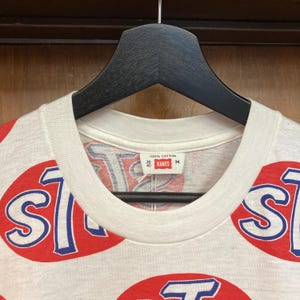 Peut inclure: Un t-shirt blanc avec un motif int&eacute;gral d'ovales rouges contenant du texte bleu et blanc. L'&eacute;tiquette int&eacute;rieure du col indique 100% Cotton, 38 40, Hanes et M. Le v&ecirc;tement est pr&eacute;sent&eacute; sur un cintre en plastique noir sur fond en bois.