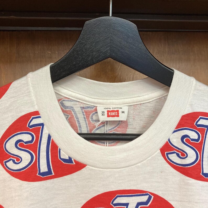 Peut inclure: Un t-shirt blanc avec un motif int&eacute;gral d'ovales rouges contenant du texte bleu et blanc. L'&eacute;tiquette int&eacute;rieure du col indique 100% Cotton, 38 40, Hanes et M. Le v&ecirc;tement est pr&eacute;sent&eacute; sur un cintre en plastique noir sur fond en bois.