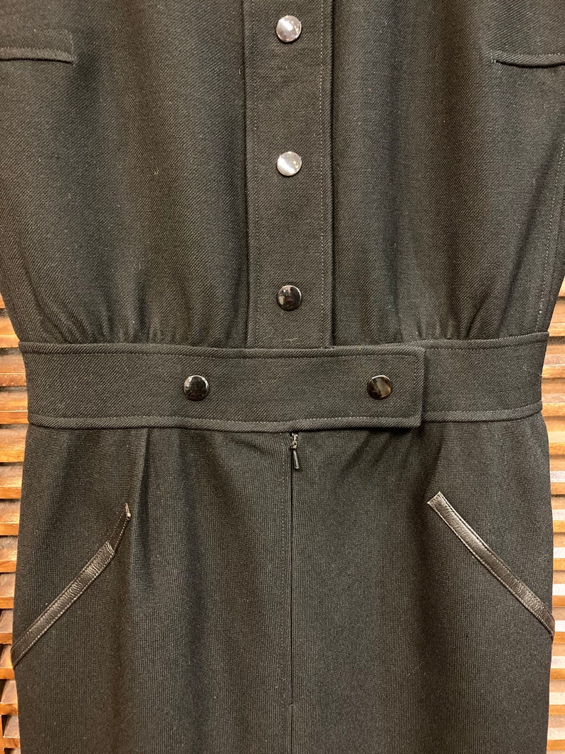 Puede incluir: Un vestido negro con cierre de botones, cintur&oacute;n y dos bolsillos con ribetes de cuero.