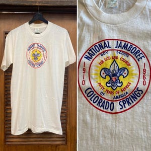 May include: Off-white t-shirt featuring a circular graphic for the National Jamboree in Colorado Springs. The graphic includes text and a Boy Scouts emblem in red, yellow, and blue. The shirt is hanging on a black hanger.
