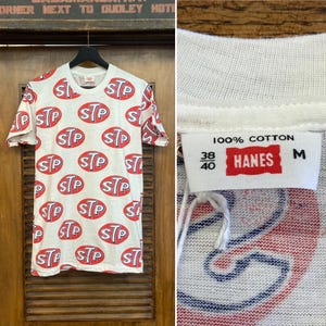 Peut inclure: Un t-shirt blanc &agrave; manches courtes avec un imprim&eacute; int&eacute;gral du logo STP en rouge et bleu. L'&eacute;tiquette int&eacute;rieure du col indique 100% coton, taille M et 38/40. Le t-shirt est pr&eacute;sent&eacute; sur un cintre noir devant un fond en bois avec du texte.