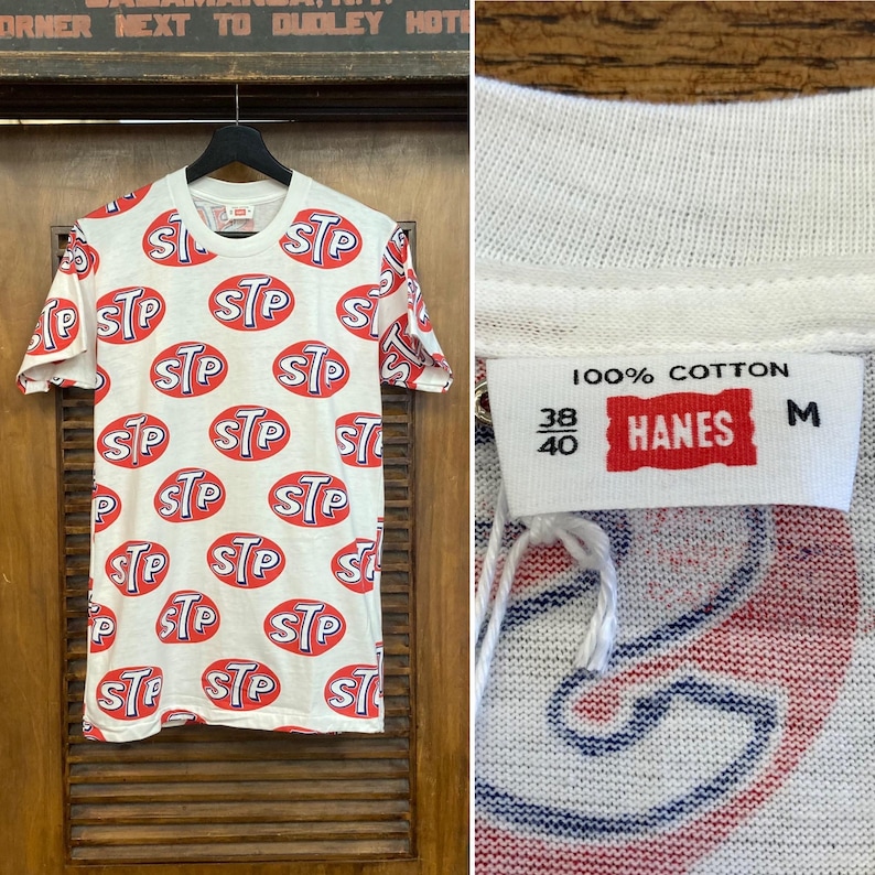 Peut inclure: Un t-shirt blanc &agrave; manches courtes avec un imprim&eacute; int&eacute;gral du logo STP en rouge et bleu. L'&eacute;tiquette int&eacute;rieure du col indique 100% coton, taille M et 38/40. Le t-shirt est pr&eacute;sent&eacute; sur un cintre noir devant un fond en bois avec du texte.