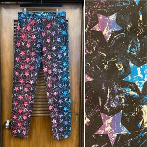 May include: A pair of black jeans with a colorful star pattern. The stars are pink, blue, and white and are scattered randomly across the fabric. The jeans are hanging on a hanger against a wooden door.