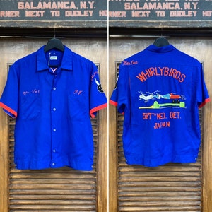 May include: A blue bowling shirt with red trim and a white button closure. The shirt has a graphic of a bowling ball and the text "CALIFO" on the left sleeve and the text "Harlan WHIRLYBIRDS 587TH MED. DET. JAPAN" on the right sleeve.