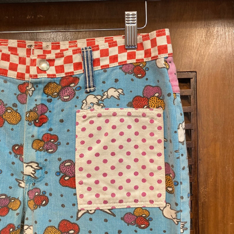 May include: Blue denim pants with a red and white checkered waistband and a pink polka dot pocket. The pants have a whimsical print of colorful balloons and clouds.