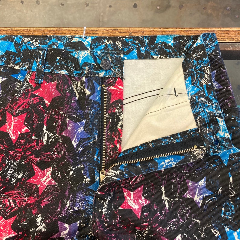 May include: Black denim jeans with a colorful star print in shades of red, blue, and purple. The jeans have a zipper fly and a button closure.