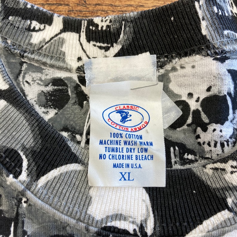 Puede incluir: Una camiseta de algod&oacute;n con estampado de calaveras en blanco y negro con una etiqueta blanca que dice "Classic Cotton Armor" e incluye instrucciones de cuidado. La etiqueta tambi&eacute;n dice "100% algod&oacute;n", "Lavar a m&aacute;quina con agua tibia", "Secar en secadora a baja temperatura", "No usar blanqueador con cloro", "Hecho en EE. UU." y "XL".
