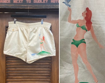 May include: White satin swim trunks with a graphic print of a woman in a swimsuit standing on a beach with a bird flying overhead. The text "SALAMANCA, N.Y." is printed on the trunks.