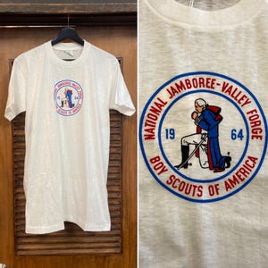 May include: White t-shirt featuring a circular graphic with the text "NATIONAL JAMBOREE-VALLEY FORGE" and "BOY SCOUTS OF AMERICA". The graphic includes a figure in blue and red attire, with the numbers "19" and "64".