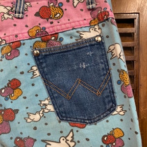May include: Blue denim jeans with a back pocket and a colorful pattern of birds and fruit on a light blue background. The pattern includes red, yellow, pink, and black.