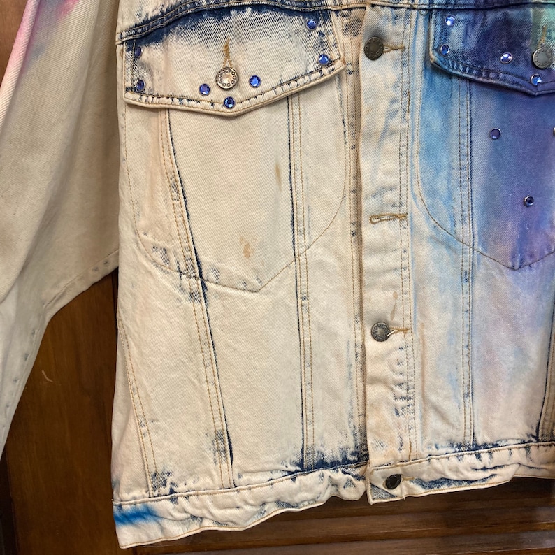 May include: A light blue denim jacket with a faded white and blue tie-dye pattern. The jacket has a pocket on the left side and a button closure. The jacket is decorated with rhinestones.