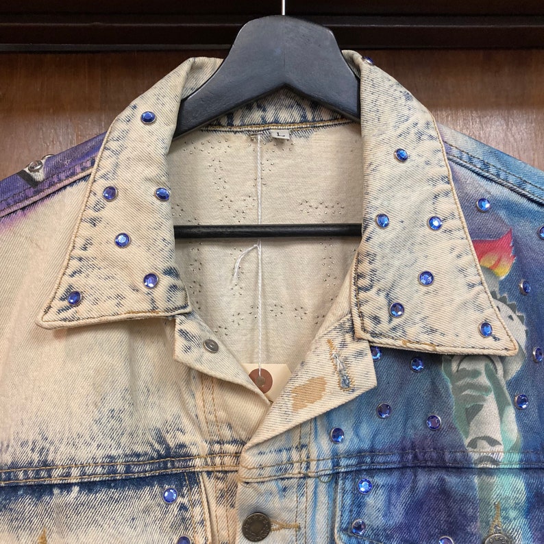 May include: A light blue denim jacket with a white and blue paint design. The jacket has a collar with blue rhinestones and a white tag on the inside.