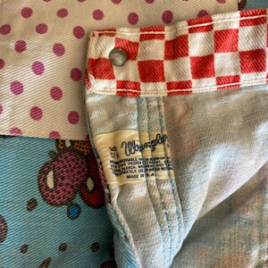 May include: A close-up of a blue denim fabric with a red and white checkered pattern. The fabric has a label that says "Wrangler" and "Size 13/4" with instructions for washing and care. The fabric is made in the USA.