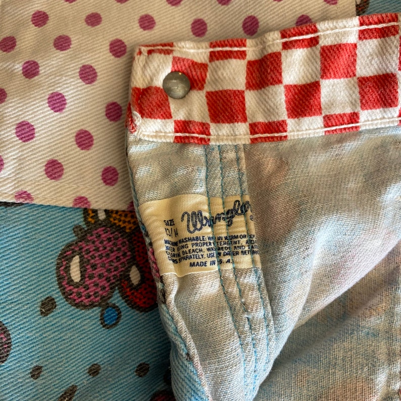 May include: A close-up of a blue denim fabric with a red and white checkered pattern. The fabric has a label that says "Wrangler" and "Size 13/4" with instructions for washing and care. The fabric is made in the USA.